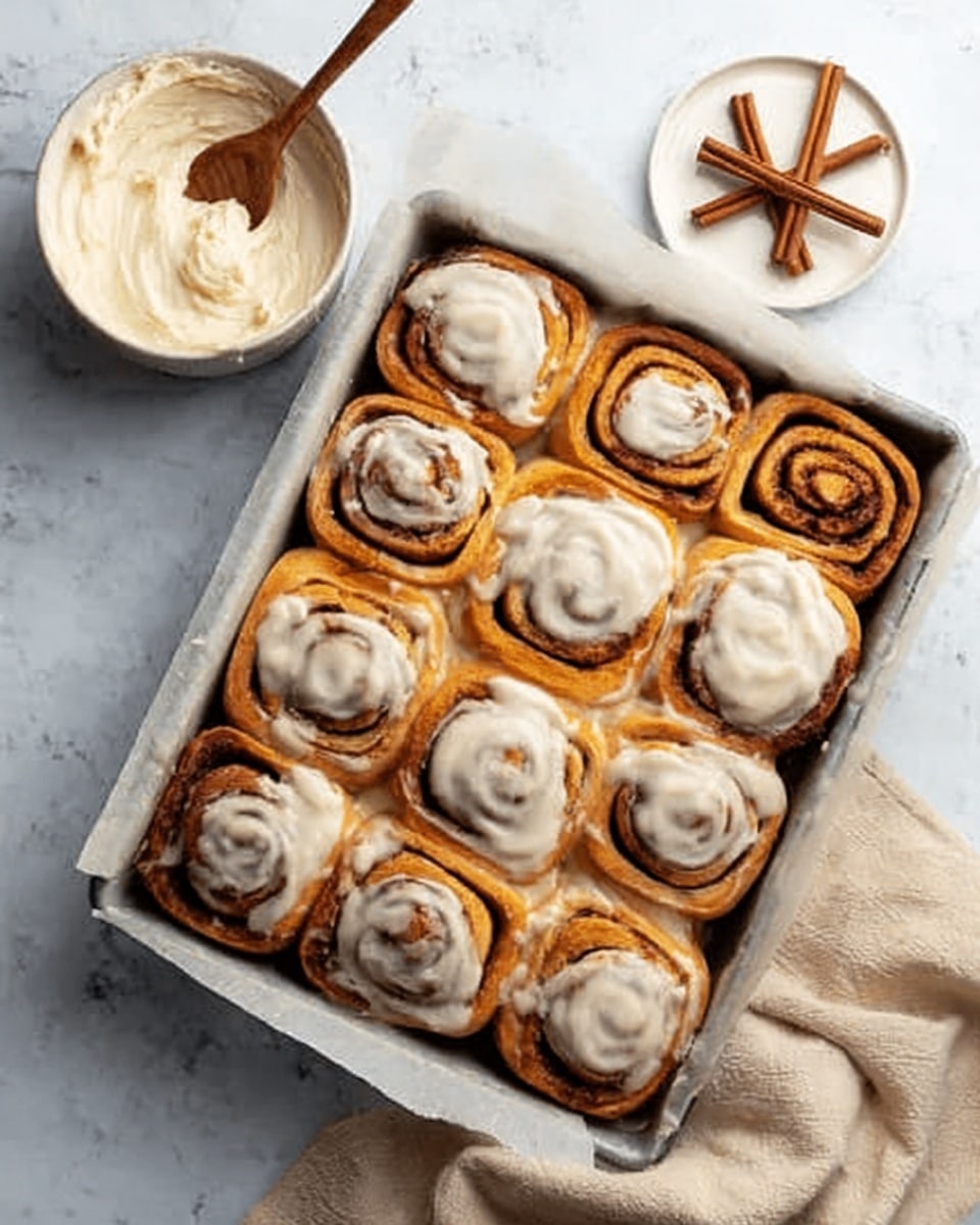 A baking tray lined with white parchment paper holds 12 cinnamon rolls arranged in three rows of four. Each roll has a golden-brown dough base with a spiral pattern of darker brown cinnamon filling visible inside. The rolls are topped with a thick, creamy white icing that spreads unevenly over the surface, some dripping down the sides slightly. To the left, there is a white bowl containing smooth cream cheese frosting with a wooden spoon resting inside it, and next to it is a small white plate with two cinnamon sticks. The whole scene is set on a white marbled surface with a soft-colored cloth partly visible in the bottom right corner. photo taken with an iphone --ar 4:5 --v 7