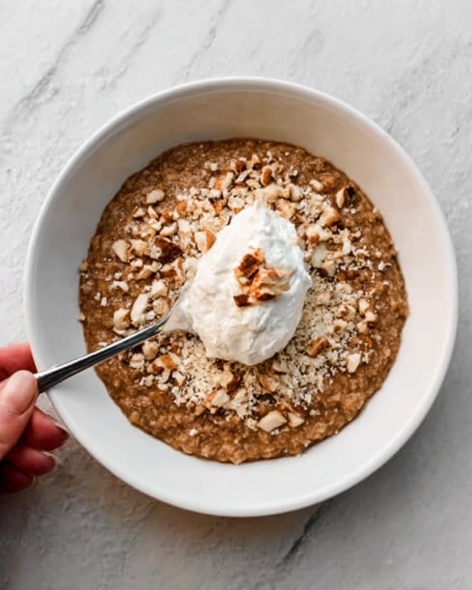 The image shows a white bowl filled with a thick brown oatmeal. On top, there is a generous layer of crushed light tan nuts scattered all over the oatmeal. In the center, a dollop of white cream with a fluffy texture sits neatly. A silver spoon is inside the bowl, resting on the left side, held by a woman's hand. The bowl is set on a surface with a white marbled texture. photo taken with an iphone --ar 4:5 --v 7