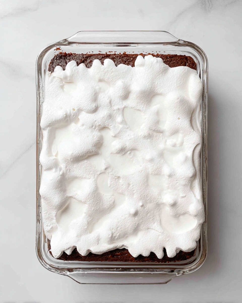 A clear rectangular glass baking dish holds a dessert with two main layers visible from the top: the bottom layer is dark brown and looks like a cooked cake, while the top layer is a thick, fluffy, white layer spread unevenly over the cake, showing some areas of the cake through small gaps. The white layer has a soft texture with small dimples scattered around. The dish is placed on a white marbled surface. photo taken with an iphone --ar 4:5 --v 7