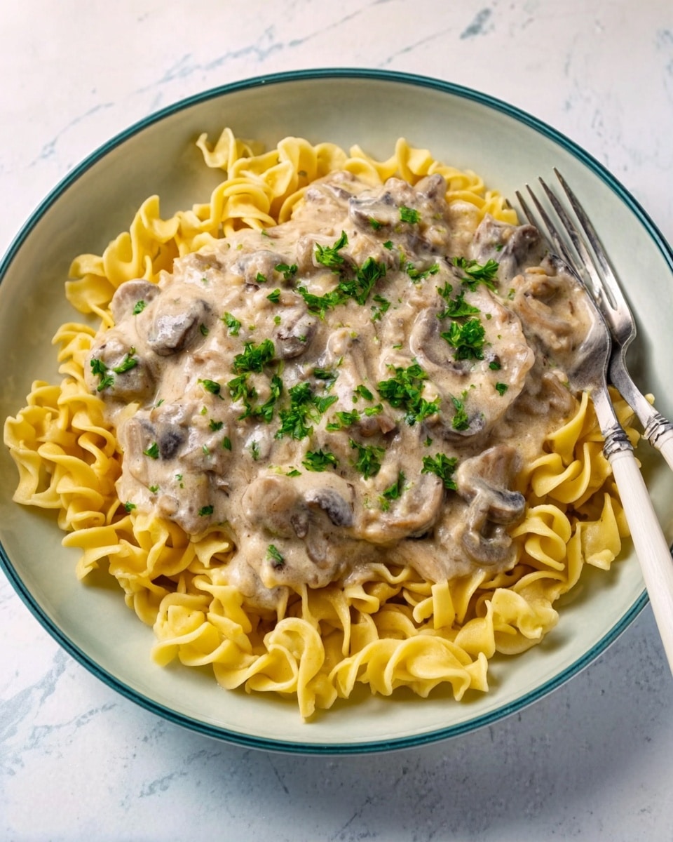 A white bowl filled with egg noodles at the bottom layer, showing a curly and yellow texture. On top of the noodles, there is a thick creamy sauce that is pale beige in color with small pieces of mushrooms scattered throughout. The dish is garnished with small green parsley leaves spread evenly over the sauce. The bowl sits on a white marbled surface, next to a white knife and fork. Photo taken with an iphone --ar 4:5 --v 7