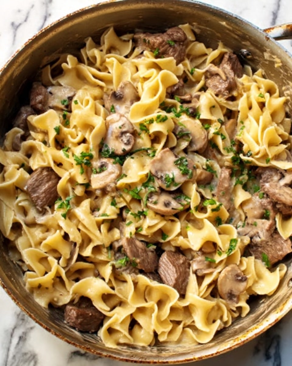 The image shows a close-up of a wide pan filled with cooked egg noodles mixed with pieces of cooked beef and small slices of mushrooms. The noodles are light yellow and look soft with a slightly ruffled texture. The beef pieces are medium brown and scattered throughout the noodles. Small bits of green herbs are sprinkled on top, adding a fresh touch. The background has a white marbled texture. Photo taken with an iphone --ar 4:5 --v 7