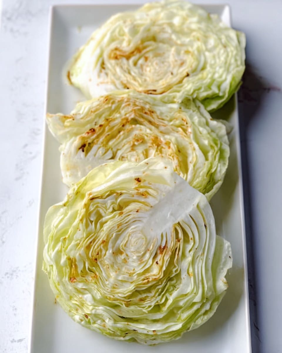 The image shows three round slices of cooked cabbage placed on a long white plate, arranged vertically. Each cabbage slice has layers of light green and pale yellow leaves visible, with a soft texture and some slight browning around the edges. The cabbage slices display natural folds and a slight translucence from cooking, sitting on a white marbled surface. A metal spatula with a silver blade rests on the plate above the top cabbage slice. Photo taken with an iphone --ar 4:5 --v 7
