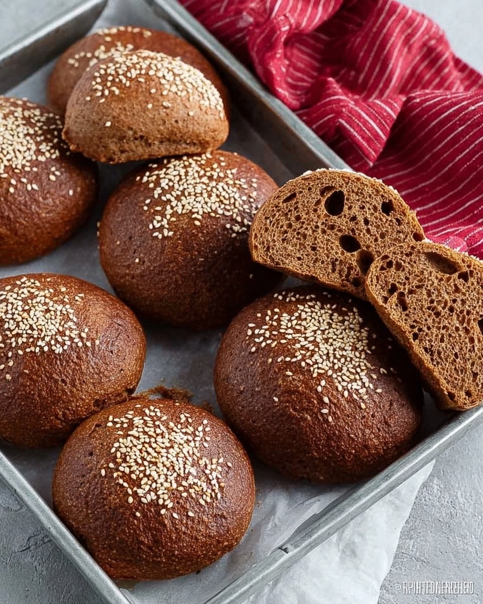 The image shows a silver baking tray with nine brown sesame seed buns arranged in two rows, some whole and some sliced in half to reveal a soft, dense, textured inside. The buns have a rough surface with sesame seeds sprinkled on top, giving a slightly crispy look. The tray is lined with a white parchment sheet, and there is a red and white striped cloth partially visible in the top right corner. The background is a white marbled surface with scattered sesame seeds around the tray. photo taken with an iphone --ar 4:5 --v 7