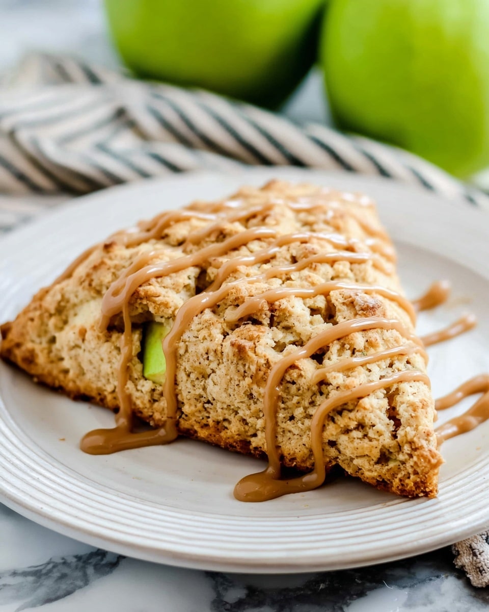 A triangular scone sits in the center of a round white plate with a ridged edge. The scone has a rough, crumbly texture with a light golden-brown color, showing small pieces of green apple embedded inside. It is topped with several thin lines of light brown glaze in a zigzag pattern across the top. The plate is placed on a white marbled surface, with a striped cloth and green apples blurred softly in the background. Photo taken with an iphone --ar 4:5 --v 7