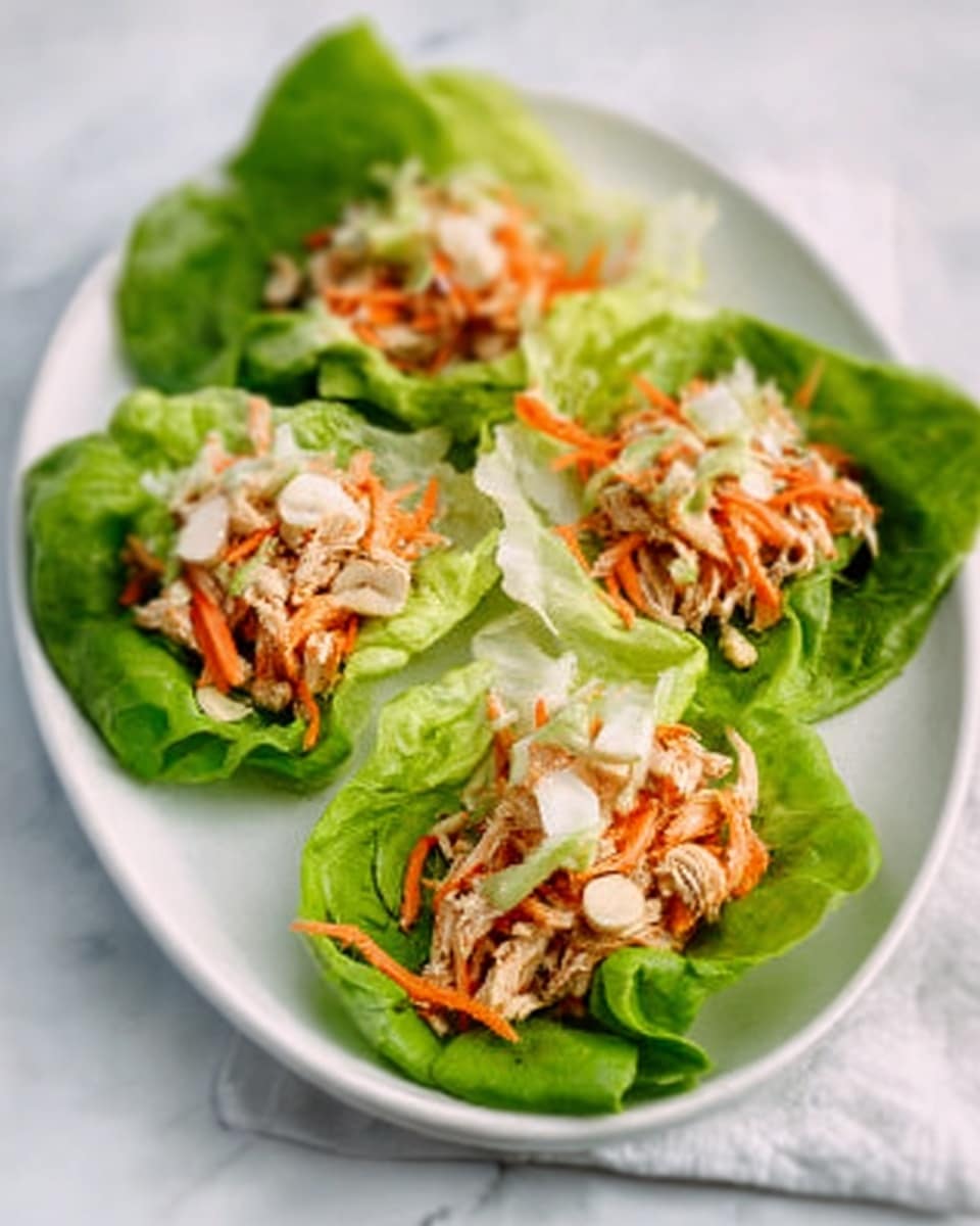 The image shows four fresh lettuce wraps placed on a white oval plate on a white marbled surface. Each wrap has a bright green lettuce leaf base, topped with a mix of shredded orange carrots, pale beige cooked chicken pieces, and small white sliced nuts or seeds. The layers create a colorful and fresh look, with the green lettuce leaves curving gently around the textured filling. photo taken with an iphone --ar 4:5 --v 7