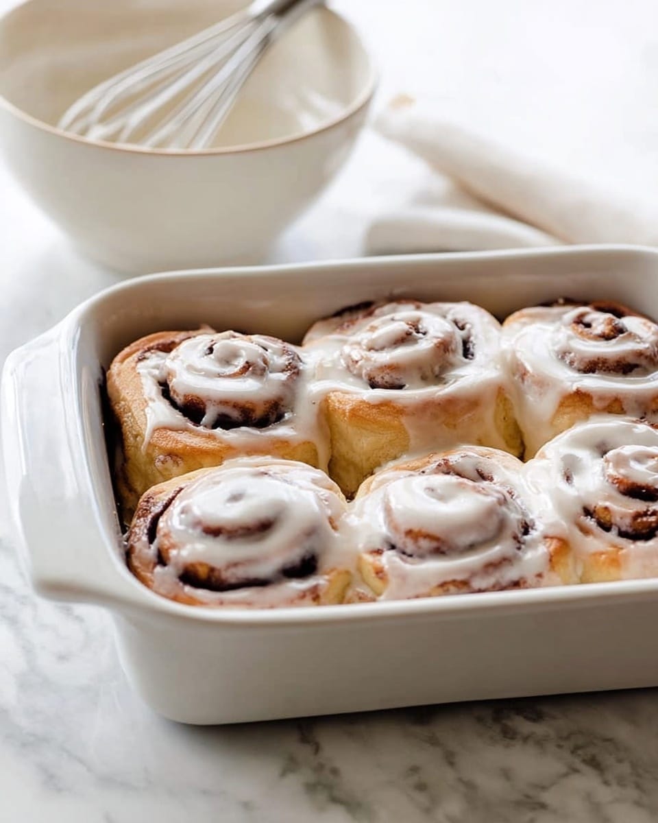 A white square baking dish holds six cinnamon rolls arranged in two rows of three. Each roll has a light brown dough with a dark brown cinnamon swirl inside, topped with a thin layer of white icing that partly melts and pools between the rolls. The rolls are soft with a slightly glossy texture from the icing. In the background, there is a white bowl with a metal whisk resting inside on a white marbled surface. Photo taken with an iphone --ar 4:5 --v 7