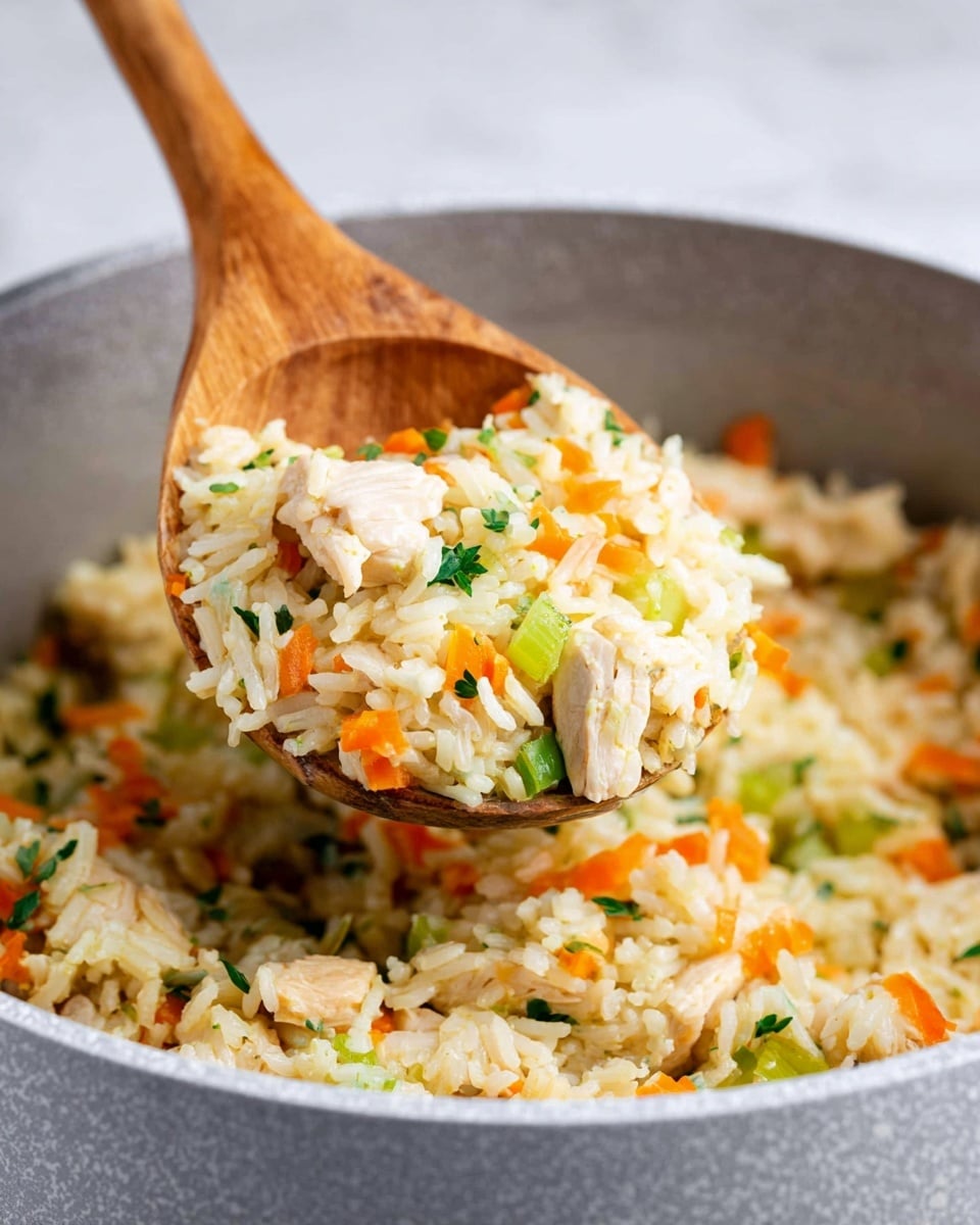 The image shows a close-up of a light gray textured pot filled with cooked yellow rice mixed with small pieces of white chicken, bright orange shredded carrots, and chopped green celery, all mixed with bits of green herbs. A wooden spoon scoops up a portion of the rice mixture, lifting it above the pot to reveal the layered mixture of colors and textures. The white marbled surface under the pot is softly blurred in the background. photo taken with an iphone --ar 4:5 --v 7