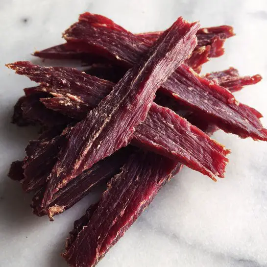 The image shows several thin strips of dried meat arranged loosely on a white marbled surface. The strips have a rich reddish-brown color with a slightly rough texture and some jagged edges. They vary in size but are mostly long and narrow. The lighting highlights the dryness and firmness of the meat pieces. photo taken with an iphone --ar 4:5 --v 7
