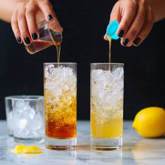 The image shows two side-by-side tall frosted glasses filled with clear ice cubes on a white marbled surface. On the left, a woman's hand with black nail polish pours a dark amber liquid from a small clear measuring cup into the glass, which has a hint of pale yellow liquid at the bottom. On the right, the same woman's hand pours a clear liquid from another small measuring cup with a blue lid into a matching glass, which has a light golden liquid surrounding the ice. Next to each glass is a bright yellow whole lemon with a small lemon peel piece placed nearby. In the background, there are short clear glasses filled with ice cubes. The background is dark, making the bright colors stand out clearly. photo taken with an iphone --ar 4:5 --v 7