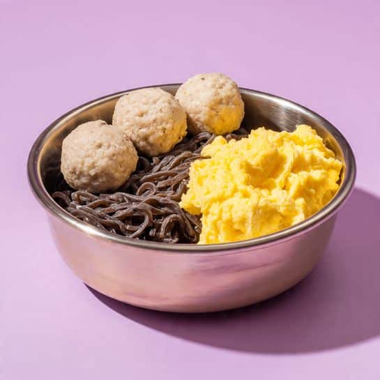 A round metal bowl with a white marbled background holds a meal with three main parts. On the left side, there are four light brown meatballs arranged in a vertical line. The center is filled with dark brown noodles that look soft and well cooked, slightly twisted and piled together. On the right side, there is a bright yellow scrambled egg with a slightly uneven texture, covering about a third of the bowl. The overall presentation is simple with clear separation between the three layers. Photo taken with an iphone --ar 4:5 --v 7