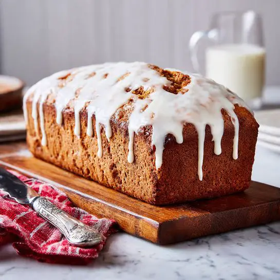 A loaf-shaped cake with a golden brown crust is placed on a wooden board on a white marbled surface. The cake has thick white icing drizzled unevenly over the top, slowly dripping down the sides in thin layers. Next to the cake is a silver knife with an ornate handle resting on a red and white checkered cloth. In the background, a glass of milk is faintly visible, slightly blurred. photo taken with an iphone --ar 4:5 --v 7