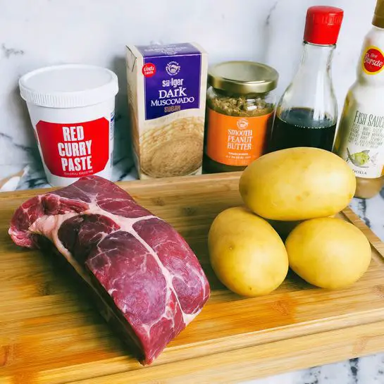 The image shows a wooden board with a raw piece of rolled red meat with visible fat on the left side, next to three whole yellow potatoes arranged in a cluster on the right. Behind the meat and potatoes, there are several containers arranged in a row: a white tub labeled