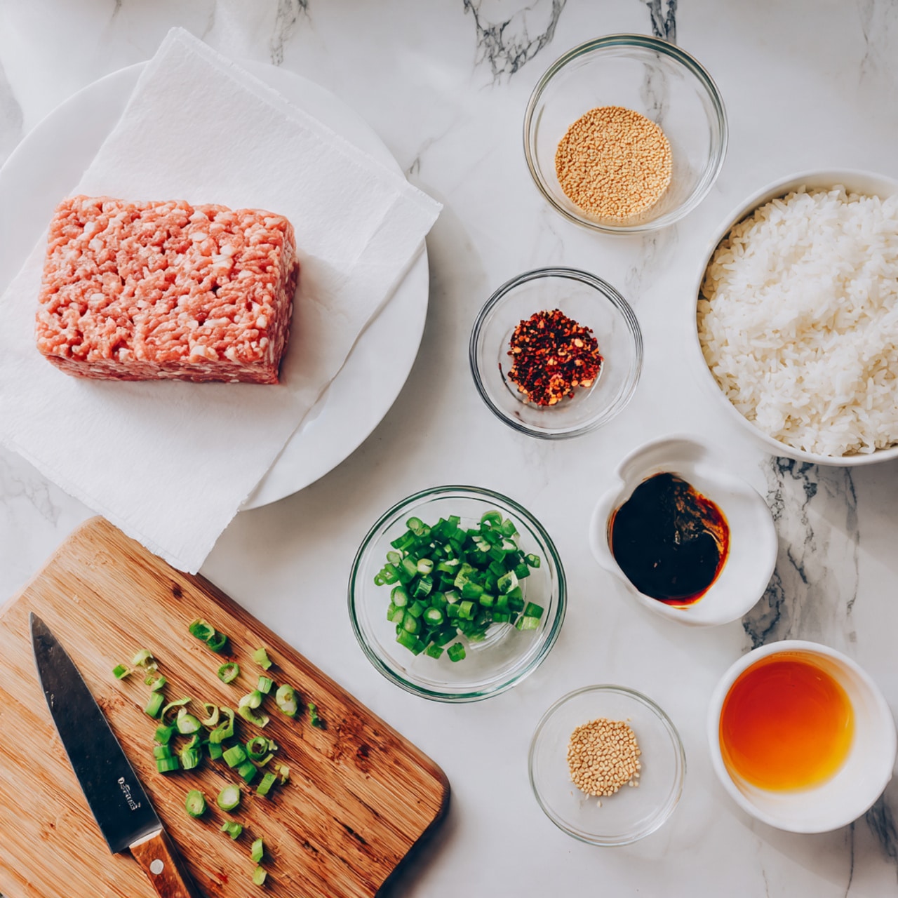 The image shows a white plate on the left with a rectangular block of raw ground meat that has a pinkish-red color and a wavy texture. To the right, there is a white bowl filled with cooked white rice, showing separate grains with a soft texture. Below the rice, there are three small white bowls containing sauces: one amber-colored, one dark soy-colored, and a clear bowl with red chili flakes. In the center bottom, a wooden cutting board holds chopped green onions scattered beside a small knife with a wooden handle. Also visible are small bowls of crushed ginger powder and toasted sesame seeds. All items are arranged on a white marbled surface. Photo taken with an iphone --ar 4:5 --v 7