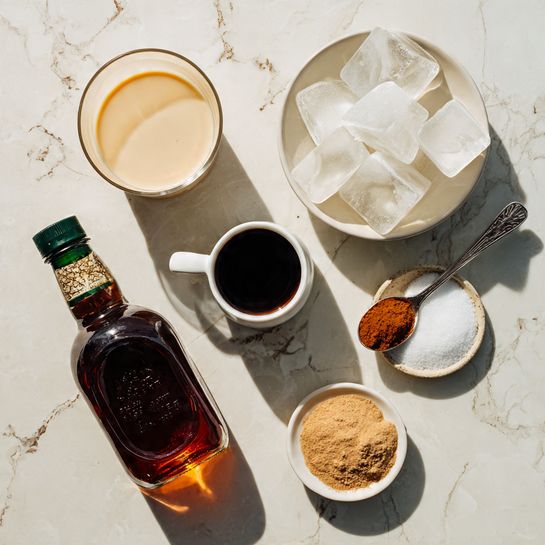 The image shows six ingredients arranged on a white marbled surface: a glass cup with light beige milk in the top left, a white bowl filled with large clear ice cubes in the top right, a small white cup with dark espresso in the center, a metal measuring spoon with dark amber maple syrup in the bottom left, a tall brown bottle of vanilla extract with a green cap below the maple syrup, and a small white bowl containing a small pile of light brown cinnamon and a small pile of white sea salt in the bottom right. Each ingredient is labeled with black text on white backgrounds. Photo taken with an iphone --ar 4:5 --v 7