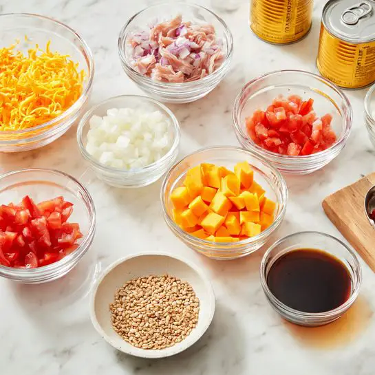 The image shows many clear glass bowls and two cans on a white marbled surface. In the front, there is a small round bowl filled with beige seeds, next to a small amount of dark brown liquid in another small bowl. Behind these are medium-sized bowls: one with small white pieces of chopped onion, another with bright orange cheese cubes, and one with finely shredded orange cheese. Toward the back, a large bowl holds shredded pale pink meat, and a medium bowl contains small red diced tomatoes. Two open cans with light yellow contents sit near the bowls. The setting is clean and bright, with everything neatly arranged for cooking. photo taken with an iphone --ar 4:5 --v 7