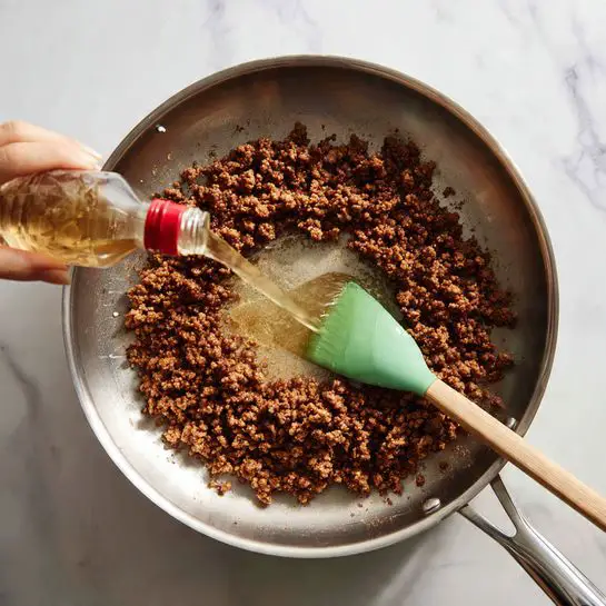 A close-up image shows a stainless steel pan on a white stove with browned, finely crumbled meat spread evenly in a ring around the pan’s sides, leaving the center mostly clear. A green spatula with a wooden handle rests in the pan on the right side among the meat. A woman's hand pours a light brown liquid from a clear bottle with a red cap into the middle of the pan where the meat is not present. The background surface is white marbled. photo taken with an iphone --ar 4:5 --v 7