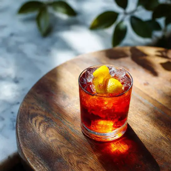 A clear glass filled with a deep orange-red liquid and ice cubes is placed on a round wooden surface. Two bright yellow lemon peels float on top of the drink, adding contrast and texture. The sunlight shines through the glass, casting a warm orange shadow on the wooden surface. The background has a white marbled texture with a few green leaves peeking into the frame at the top right corner. photo taken with an iphone --ar 4:5 --v 7
