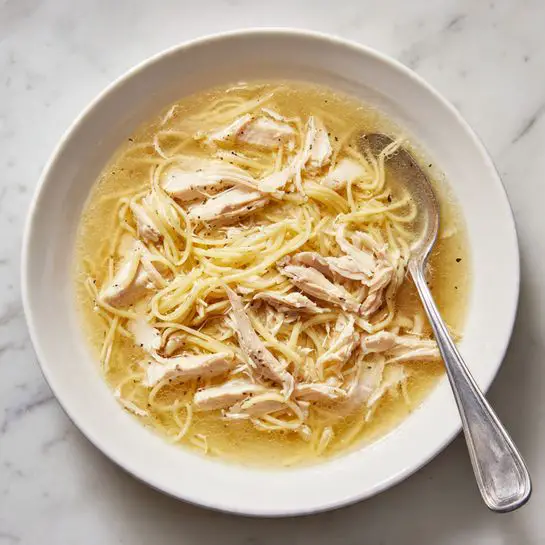 A white bowl sits on a white marbled surface filled with a dish of shredded chicken and thin noodles mixed in a light brown broth or sauce. The shredded chicken pieces are off-white and soft looking, layered over the noodles, which are thin and tangled in a similar light brown color. A metal spoon rests inside the bowl on the right side, partially submerged in the dish. The scene looks casual and warm. photo taken with an iphone --ar 4:5 --v 7