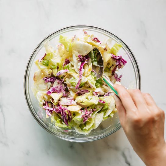 A clear glass bowl filled with a mixed salad that has creamy, light yellow dressing with visible bits of purple cabbage and pale green celery mixed throughout. A woman's hand holds the side of the bowl while a silver spoon with a slight green tint is stirring the mixture. The bowl is placed against a white marbled surface with soft natural light shining from the back. photo taken with an iphone --ar 4:5 --v 7