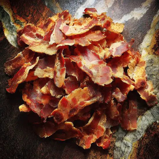 The image shows a large pile of sliced cooked meat spread out over a dark wooden surface. The meat has a mix of reddish-brown and pink colors, with some parts showing a bit of fat and crispy edges. The slices vary in thickness and are layered unevenly, giving a rough texture look. The background has a white marbled texture. The photo taken with an iphone --ar 4:5 --v 7