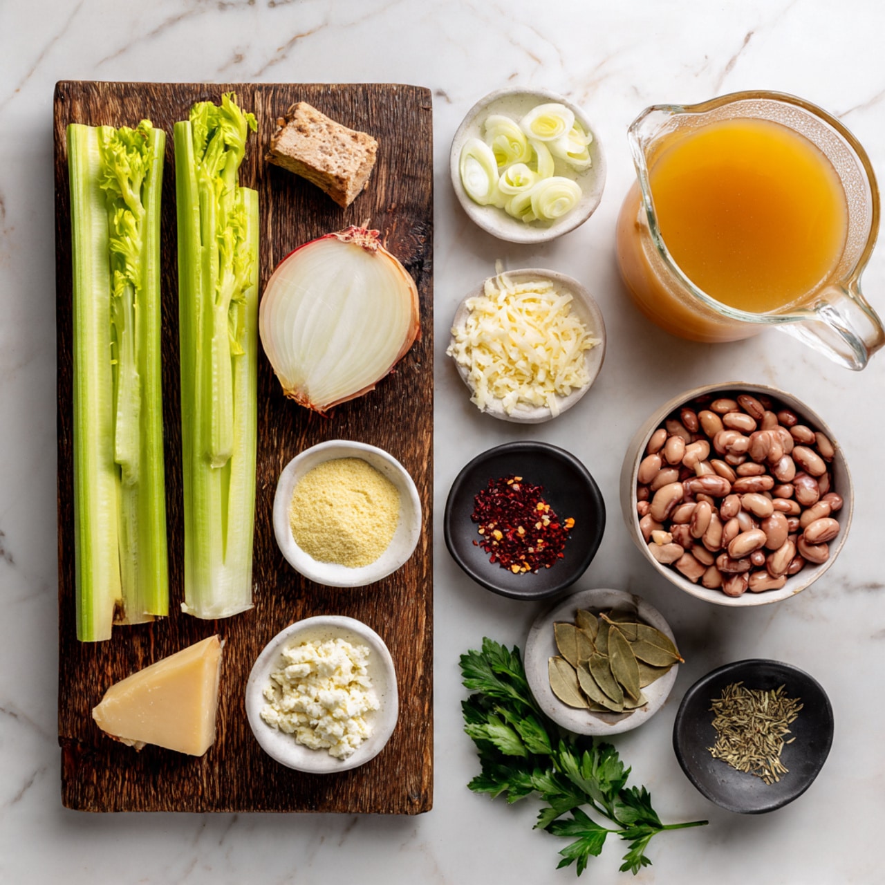 The image shows the ingredients for a recipe spread neatly on a white marbled surface. On the left, a dark wood cutting board holds three long, bright green celery stalks, a halved onion with the white inside and reddish skin, and a small piece of light brown cheese. Surrounding the board are small white bowls and black dishes holding various spices and ingredients: pale yellow cheese powder, peeled garlic cloves, dried herbs, red chili flakes, and lemon zest. To the top right, a white bowl is full of light brown kidney beans. A glass jug filled with orange-colored broth sits on the right side. Small fresh green parsley leaves add a touch of color near the edges. photo taken with an iphone --ar 4:5 --v 7