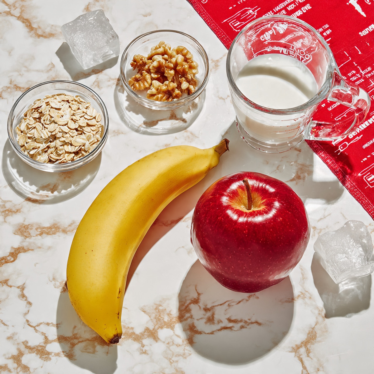 A bright yellow banana curves gently beside a smooth red apple, both placed on a white marbled surface. Around them are small clear glass bowls holding light rolled oats, golden honey, rough walnut pieces, and white ice cubes. A larger clear glass measuring cup filled with white milk sits nearby. A vibrant red cloth with white writing peeks in from the top right corner of the image. The whole setup is clean, simple, and neatly arranged in a flat lay style, photo taken with an iphone --ar 4:5 --v 7