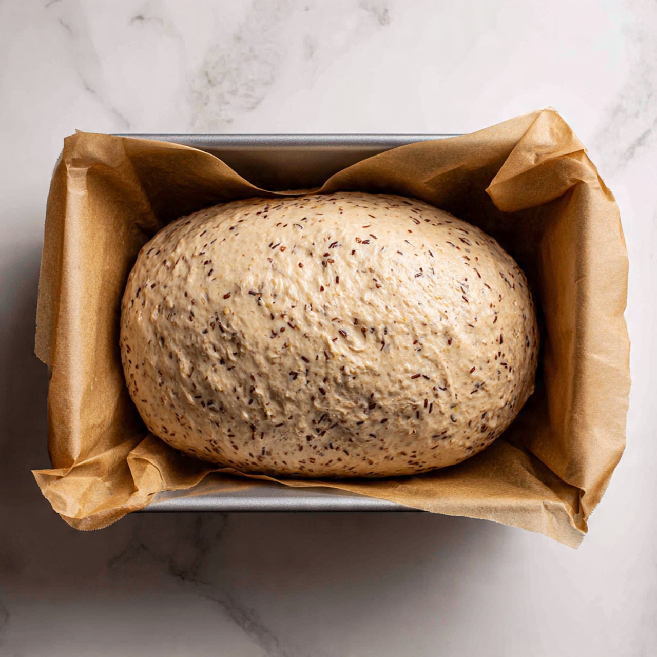 The image shows an uncooked loaf of bread dough placed inside a rectangular baking pan lined with parchment paper. The dough is oval-shaped and has a light beige color mixed with visible small dark specks and grains throughout, giving it a textured look. The surface of the dough is smooth but uneven, with some slight bulges. The baking pan is placed on a white marbled surface. photo taken with an iphone --ar 4:5 --v 7