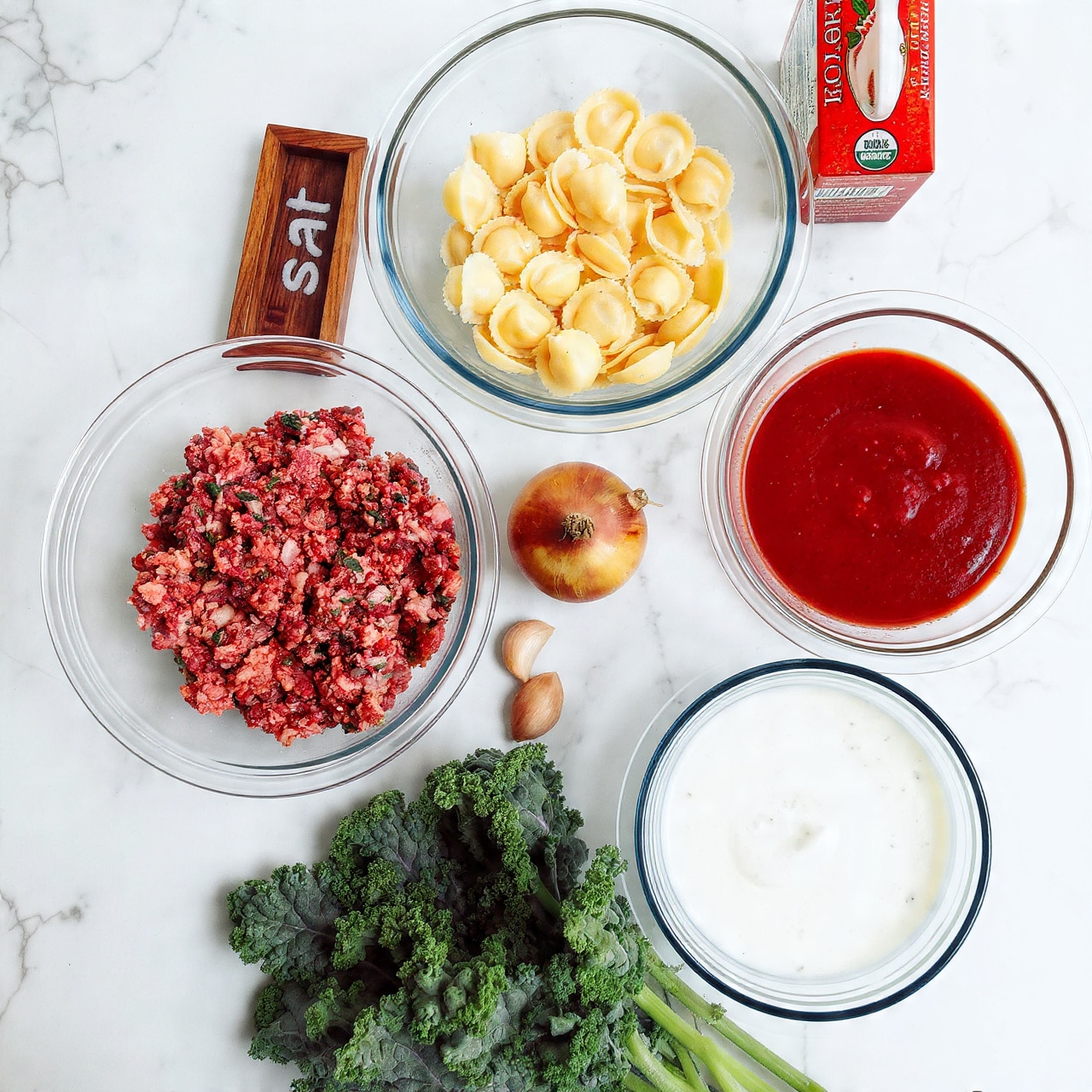 The image shows several clear glass bowls and containers arranged on a white marbled surface. One clear glass bowl holds a large amount of ground meat mixed with herbs and spices, showing a pinkish-brown color with flecks of green. Another clear glass bowl contains a pile of uncooked tortellini pasta, pale yellow with smooth, folded shapes stacked together. A third clear bowl holds a thick, bright red tomato sauce. Next to these, there is a white bowl with a blue pattern around the rim filled with a creamy white sauce. A small wooden container labeled