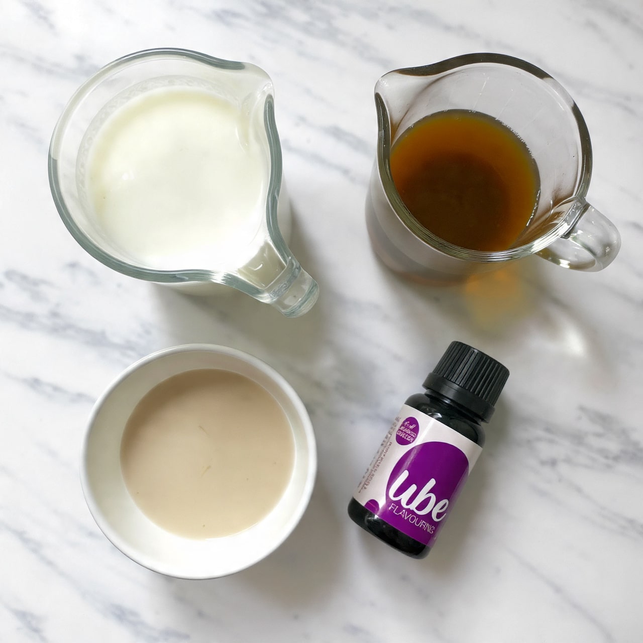 The image shows four items placed on a white marbled surface arranged in a loose square shape: a clear glass jug filled with white milk in the top left, a smaller clear glass jug with dark brown liquid in the top right, a small white bowl containing a light beige liquid in the bottom left, and a small black plastic bottle with a purple and white label that reads