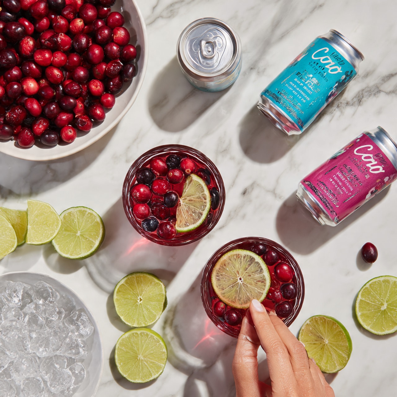 The image shows a white bowl full of whole red cranberries in the top left corner. To the right, there are two silver cans with colorful labels, one blue and one pink. Below the cranberry bowl, there are two clear glasses with red drinks inside; one has slices of lime and berries floating on top, and the other is just the red drink without any toppings. Around the glasses, there are several lime slices and wedges scattered on a white marbled surface. Near the bottom, a white bowl is filled with large clear ice cubes. A woman's hand is seen touching a lime wedge on the side. photo taken with an iphone --ar 4:5 --v 7