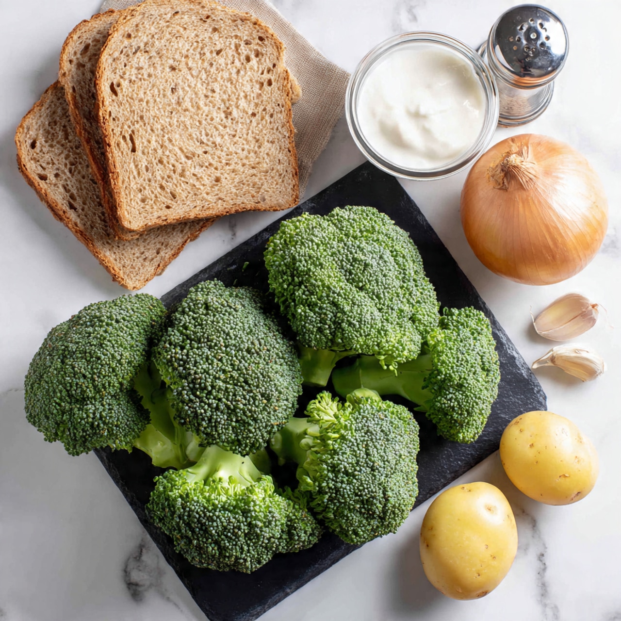A black cutting board holds several fresh broccoli florets arranged in a cluster at the center, their bright green color and textured small buds clearly visible. To the upper right, there is a small onion with a light brown skin. At the top center, a silver grinder jar stands behind a glass bowl filled with white cream. To the left, two slices of whole wheat bread rest side by side. At the bottom right, three peeled cloves of garlic and a single yellow potato sit next to the broccoli, all positioned on a clean white marbled surface. Photo taken with an iphone --ar 4:5 --v 7