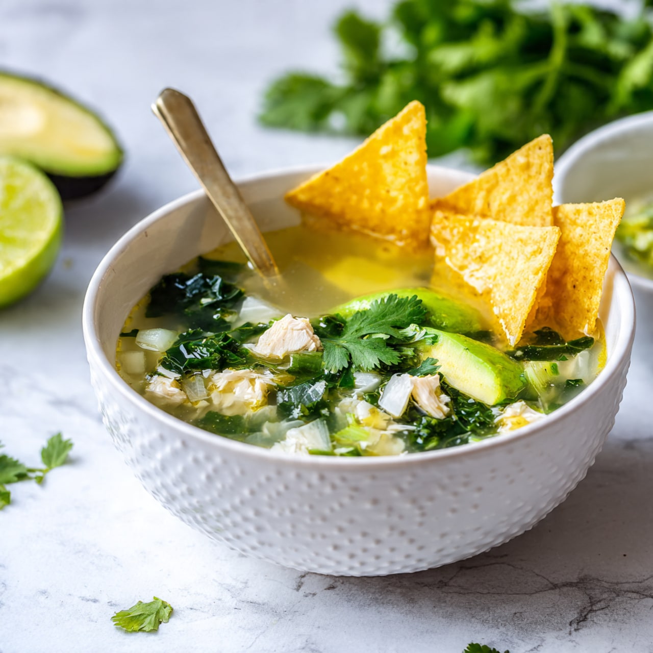 A white bowl with a honeycomb texture is filled with clear yellow broth and visible layers of small white cubes of chicken and bright green kale pieces mixed in. On the right side of the bowl, there are two large triangular, light yellow tortilla chips standing upright, next to a fresh green avocado slice with smooth texture and a sprig of bright green cilantro resting on top. A pale gold spoon is placed inside the bowl on the left side, partly submerged in the soup. In the background on the right, part of a white bowl holding more soup with similar ingredients is visible, alongside a twisted half of a squeezed lime with green skin on the white marbled surface under the bowls. Fresh cilantro sprigs and half an avocado sit in the top left of the image. photo taken with an iphone --ar 4:5 --v 7