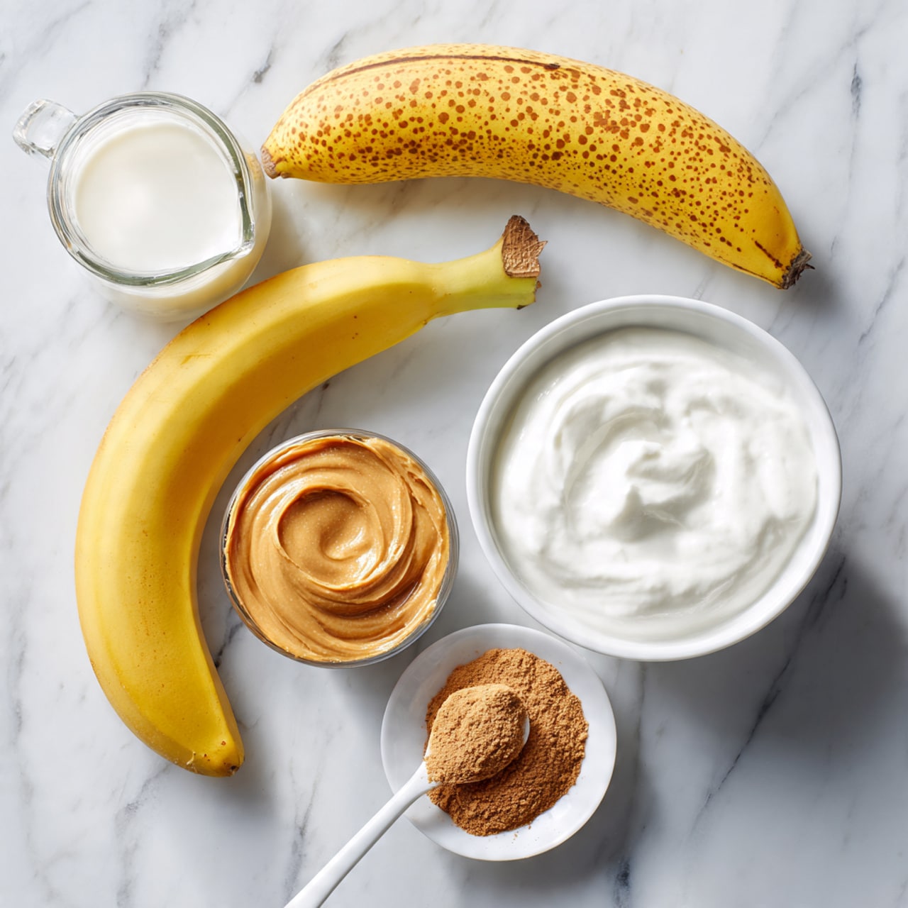 A ripe yellow banana with brown spots lies on a white marbled surface next to a clear glass jug filled with white milk. To the right, there is a white bowl filled with thick, white yogurt, and below it, a small white bowl containing fine brown powder. At the bottom left, a small white plate holds a dollop of light brown peanut butter with a white spoon resting next to it. The items are arranged neatly in a simple, clean layout. photo taken with an iphone --ar 4:5 --v 7