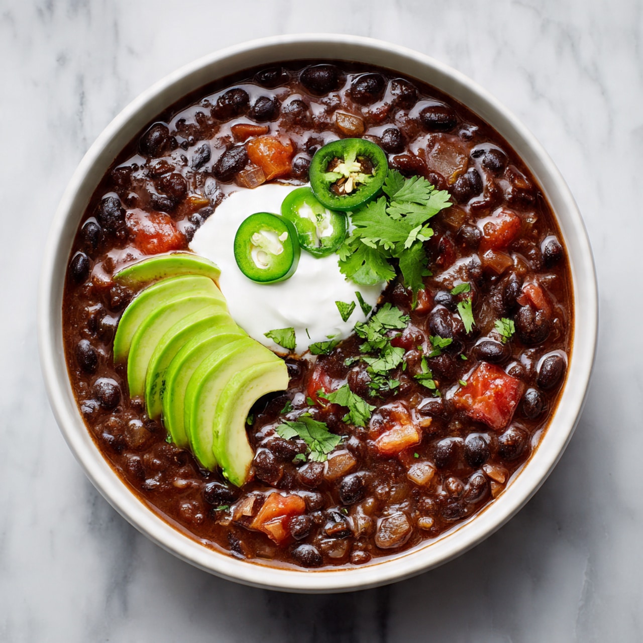 A white bowl filled with a thick black bean soup, with visible whole black beans, diced tomatoes, and cooked onions. On top, there is a round dollop of white sour cream placed slightly off center, garnished with two slices of green jalapeño and small green cilantro leaves spread around. To the side of the sour cream, there are three slices of fresh avocado arranged neatly. The bowl is placed on a white marbled surface. photo taken with an iphone --ar 4:5 --v 7