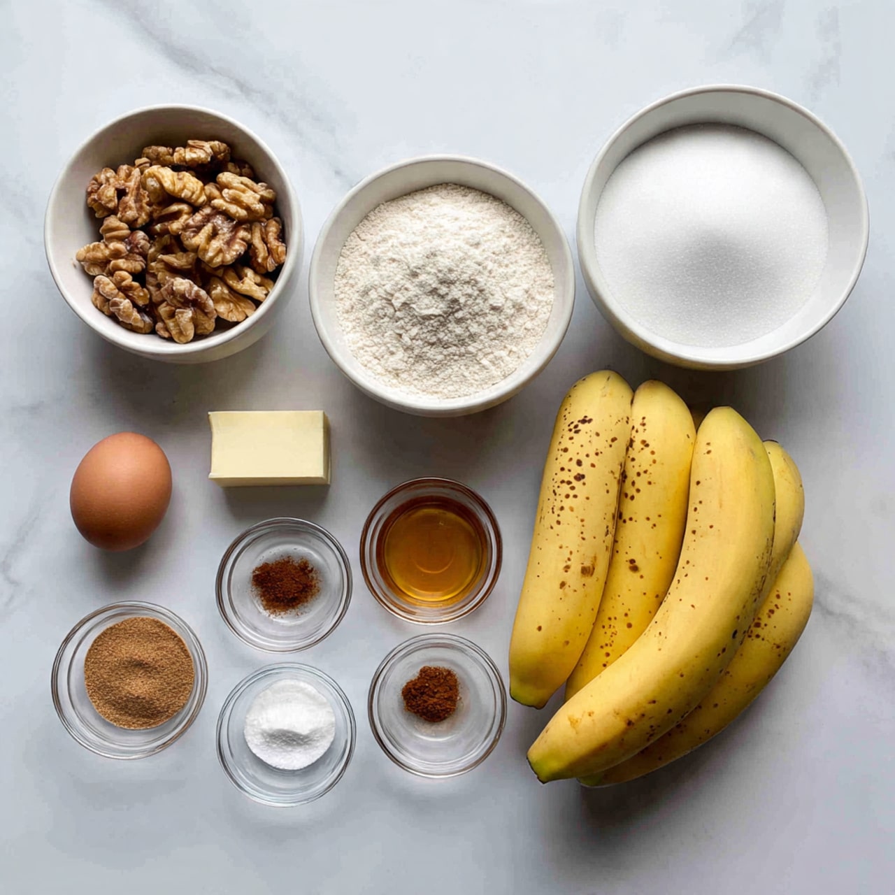 The image shows ingredients arranged on a white marbled surface: there are three ripe yellow bananas with brown spots on the right, and above them a small white bowl filled with white sugar; to the left of the sugar bowl is a white bowl with white flour, and below the flour a single brown egg and a small stick of butter. On the far left, a white bowl holds many walnut halves. Below the walnuts are four small clear glass bowls arranged in a square, containing cinnamon powder, salt, baking soda, and baking powder, with a small bowl of honey in the front center. photo taken with an iphone --ar 4:5 --v 7