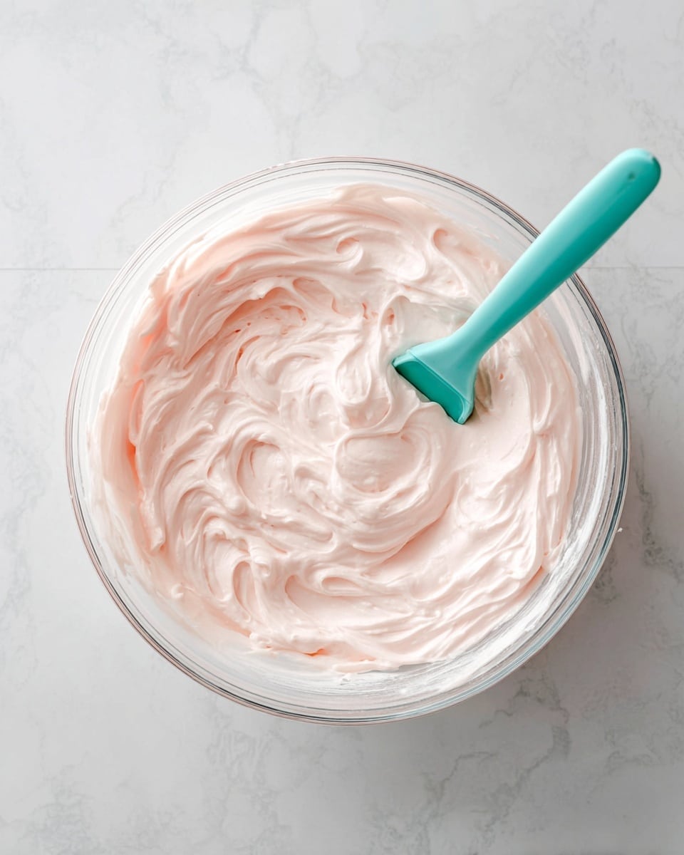 A clear glass bowl holds one thick and creamy layer of pale pink whipped frosting with smooth, soft swirls throughout. Inside the bowl, a teal silicone spatula is partially dipped into the frosting, resting angled toward the right side. The bowl is placed on a white marbled surface, which adds a subtle textured background to the image. Photo taken with an iphone --ar 4:5 --v 7