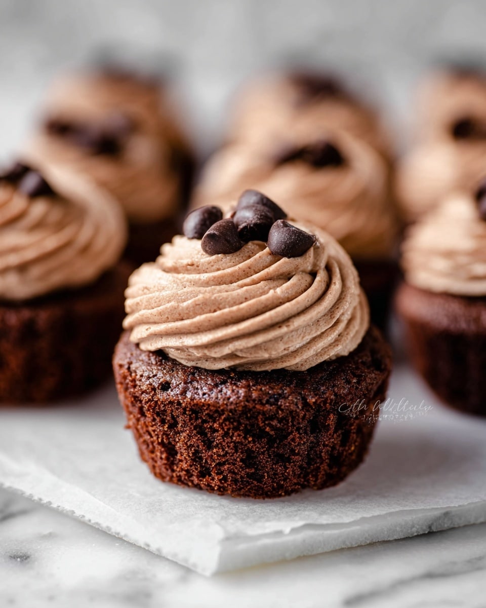 The image shows a close-up of a small chocolate cupcake with three main layers. The bottom layer is a rich, dark brown cake with a soft, slightly cracked texture. Above it, a thick swirl of light brown, smooth chocolate frosting is piped in a ridged pattern. On top of the frosting, there are several small dark chocolate curls positioned in the center. The cupcake rests on white parchment paper with more cupcakes blurred in the background, all on a white marbled surface. Photo taken with an iphone --ar 4:5 --v 7