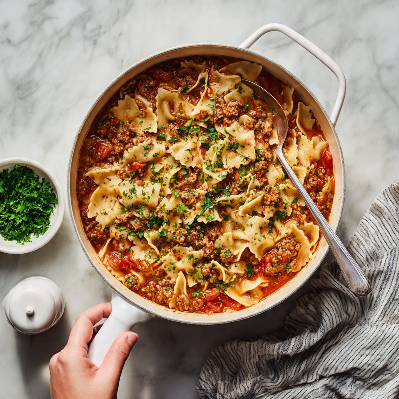 The image shows a large white pot filled with a thick, layered soup or stew. The top layer consists of flat, irregularly shaped pasta pieces, some colored creamy white and slightly soft in texture, partly covered by a sauce made of small chunks of tomatoes and ground meat in a rich reddish-orange broth. Scattered green herbs are sprinkled on top, adding a fresh touch. Next to the pot on a white marbled surface, there is a small white bowl filled with chopped fresh green herbs and a white salt grinder, with a striped cloth napkin partially visible. A woman's hand holds the pot's handle, showing a careful grip. photo taken with an iphone --ar 4:5 --v 7