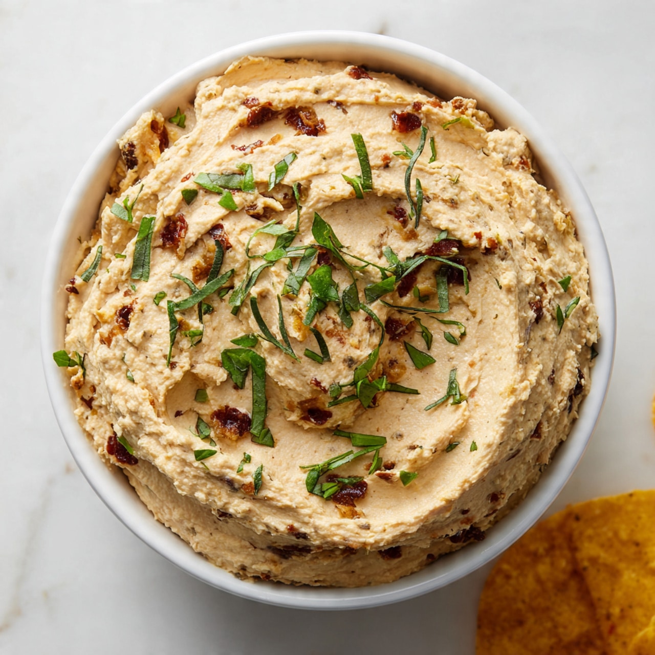 A white bowl filled with a thick, creamy beige dip that has visible small chunks mixed throughout. The dip has a smooth, slightly lumpy texture with bits of brown and lighter tones, likely from ingredients mixed in. On top, small pieces of fresh green herbs are sprinkled unevenly, adding a pop of color. The bowl sits on a white marbled surface with a small edge of a yellow chip peeking in at the bottom right. Photo taken with an iphone --ar 4:5 --v 7