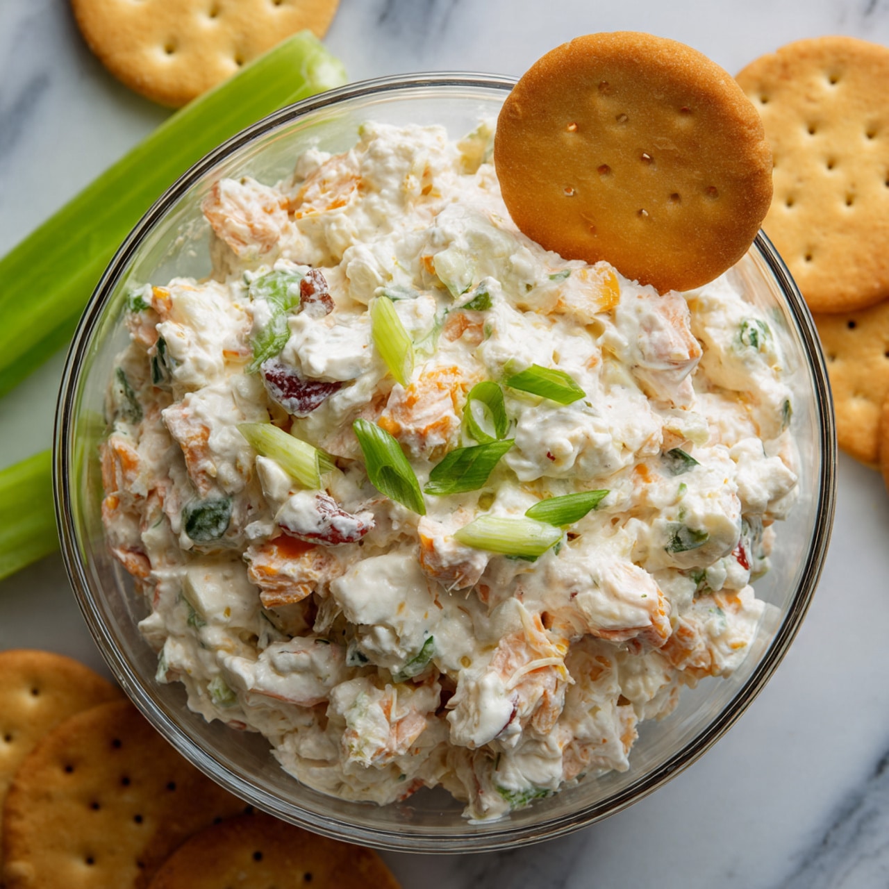 A clear round glass bowl filled with a creamy salad that has small white and pale orange chunks, mixed with bits of green herbs and red pieces. On top of the salad, there are chopped green onions scattered and a round golden cracker placed standing in the center. Around the bowl, there are more golden round crackers and a piece of green celery resting on a white marbled surface. The photo taken with an iphone --ar 4:5 --v 7