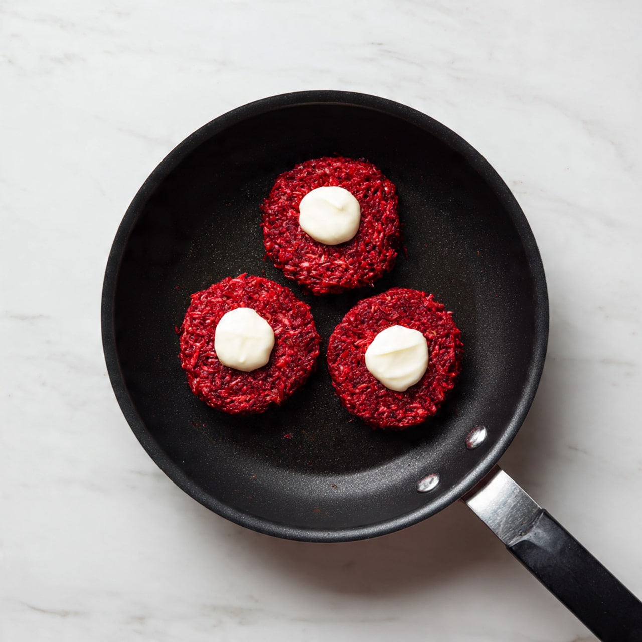 In a black frying pan set on a white marbled surface, there are three small, round red patties arranged in a triangle. Each patty is topped in the center with a dollop of white cream that stands out against the deep red texture of the patties. The patties have a slightly rough surface showing the finely shredded ingredients. The dark pan contrasts with the bright red and white colors, making the patties look fresh and vibrant. photo taken with an iphone --ar 4:5 --v 7