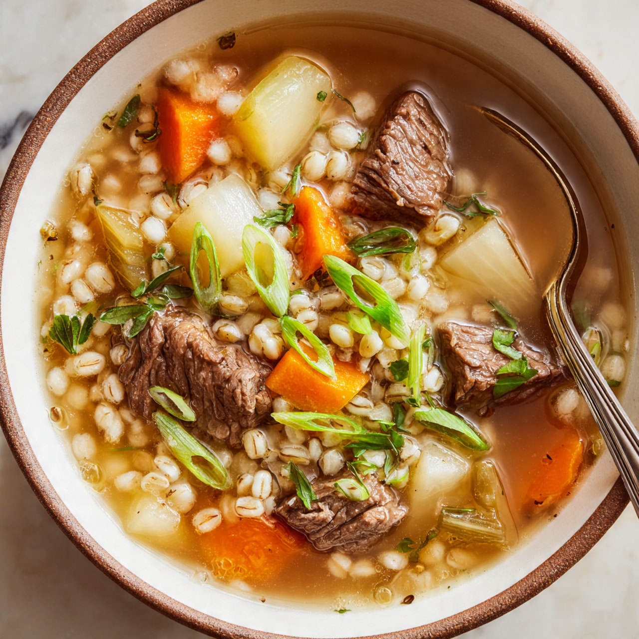 A close-up of a bowl of barley beef soup is shown, with three main layers visible: the bottom layer is a clear, light brown broth; the middle layer has chunks of tender brown beef, light beige barley grains, orange carrot slices, pale yellow potato pieces, and bits of translucent celery and onion; the top layer is scattered green herbs and thin rings of green onion as garnish. The soup is served in a white bowl with a brown rim on a white marbled surface. A silver spoon rests inside the bowl. Photo taken with an iphone --ar 4:5 --v 7