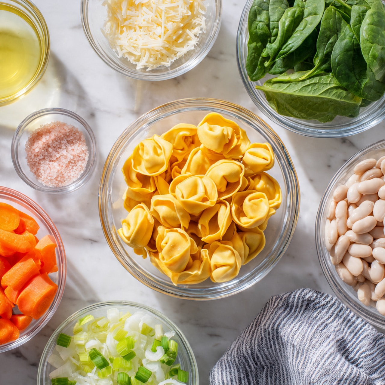 The image shows multiple clear glass bowls arranged on a white marbled surface, each bowl filled with different ingredients. In the center, there is a bowl filled with yellow tortellini pasta. Surrounding it, clockwise from the top left, there is a pale yellow liquid in a small glass, finely chopped white onion, chopped light green celery, sliced bright orange carrots, white beans, a sprig of fresh green parsley, pink salt and pepper in a small clear bowl, light yellow grated cheese, and fresh green spinach leaves. A striped cloth napkin is partially visible on the right side of the image. Photo taken with an iphone --ar 4:5 --v 7