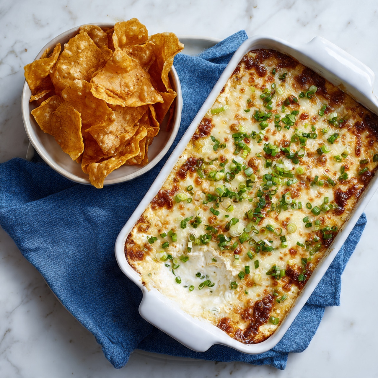 A white rectangular dish filled with a baked dip showing two main layers: a golden melted cheese layer on top dotted with finely chopped green herbs, and a white creamy layer beneath with small green onion pieces visible; a white small bowl next to it holds crisp, golden-brown wonton chips, all placed on a white marbled surface with a blue cloth partially under the dish photo taken with an iphone --ar 4:5 --v 7