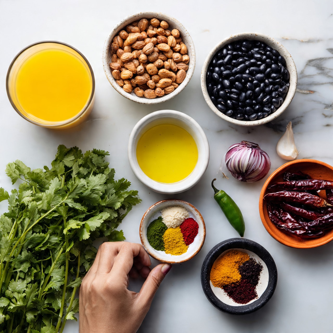 This image shows ingredients for cooking placed on a white marbled surface. From left to right and top to bottom, there is a glass of yellow juice or broth, a white bowl filled with small brown peanuts, a bunch of fresh green cilantro, a white bowl filled with black beans, a white bowl with light yellow oil, a red onion, two cloves of garlic placed next to a green chili, a small white bowl with three colorful powders (likely spices) including red, green, and yellow powders, a small orange bowl with dark red dried chili peppers, and a small black bowl containing black paste or sauce. A woman's hand is not shown but could be imagined reaching for one of the items. The photo taken with an iphone --ar 4:5 --v 7