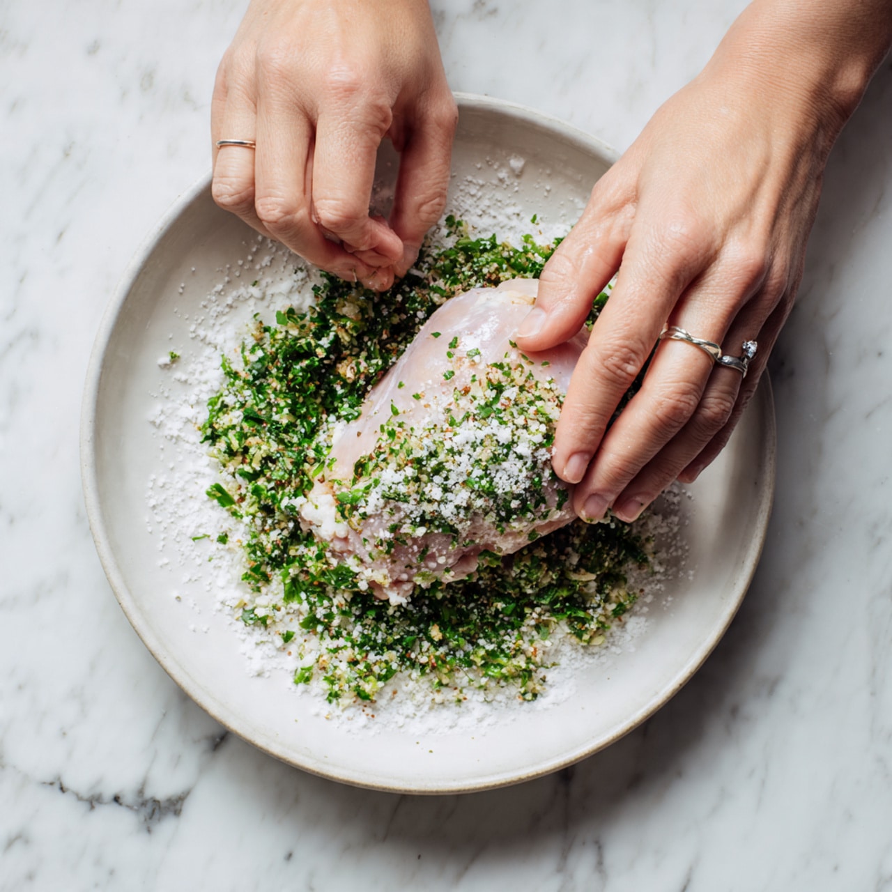 A woman's hands are holding a raw chicken piece that is coated with small white granules, likely a seasoning or salt, over a thick layer of finely chopped green herbs mixed with a white powder, all placed on a white plate. The plate is on a white marbled surface. The chicken’s texture looks smooth and moist, with the herbs providing a fresh, textured base around it. The woman's ring is visible on one finger. Photo taken with an iphone --ar 4:5 --v 7