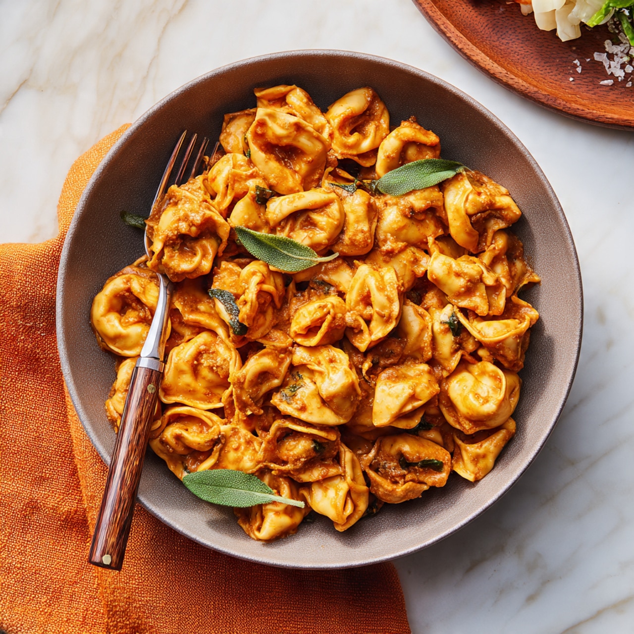 A round gray bowl filled with about two layers of tortellini pasta coated in an orange-brown sauce. The pasta pieces are slightly curled and soft-looking, guided by a few green sage leaves scattered on top for color contrast. A silver fork with a wooden handle is placed on the left side inside the bowl. The bowl rests on a white marbled textured surface with a hint of an orange cloth in the bottom left corner and a partially visible round wooden board with food in the upper right corner. Photo taken with an iphone --ar 4:5 --v 7
