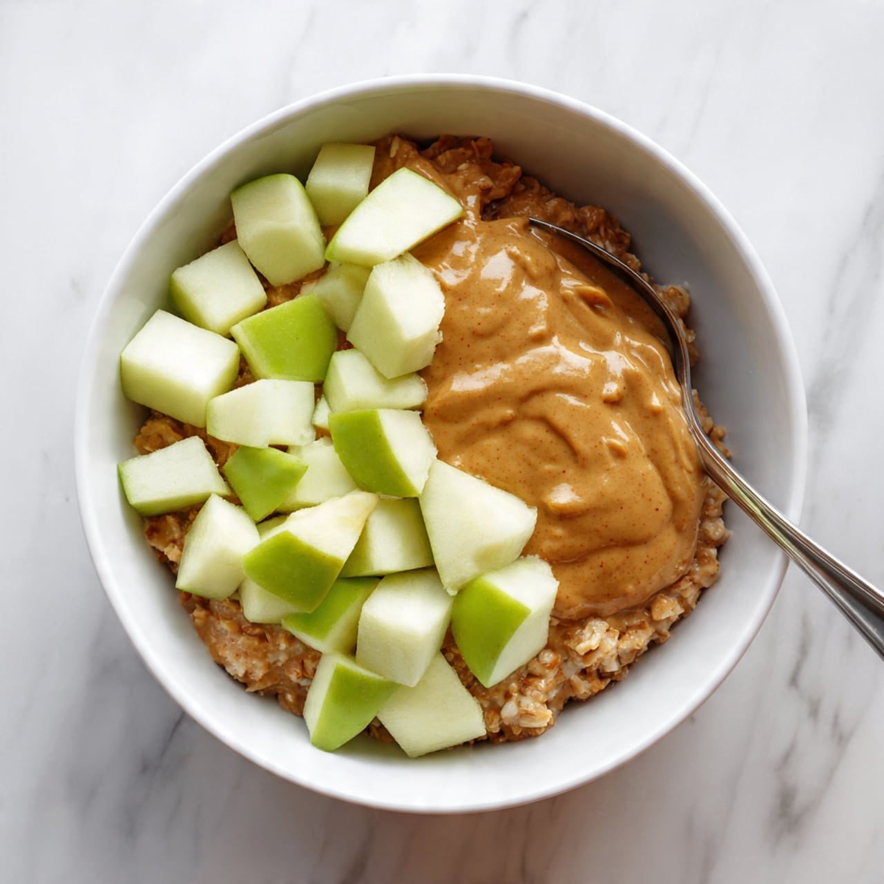 A white bowl filled with a base layer of oatmeal that has a rough, soft texture. On top of the oatmeal, there is a thick layer of light brown creamy peanut butter sauce spreading evenly but a bit irregularly. Scattered on the top is a layer of green apple chunks with white inside, some covered partly by the peanut butter sauce. A metal spoon is seen partially inside the bowl near the side. The bowl sits on a white marbled surface. Photo taken with an iphone --ar 4:5 --v 7