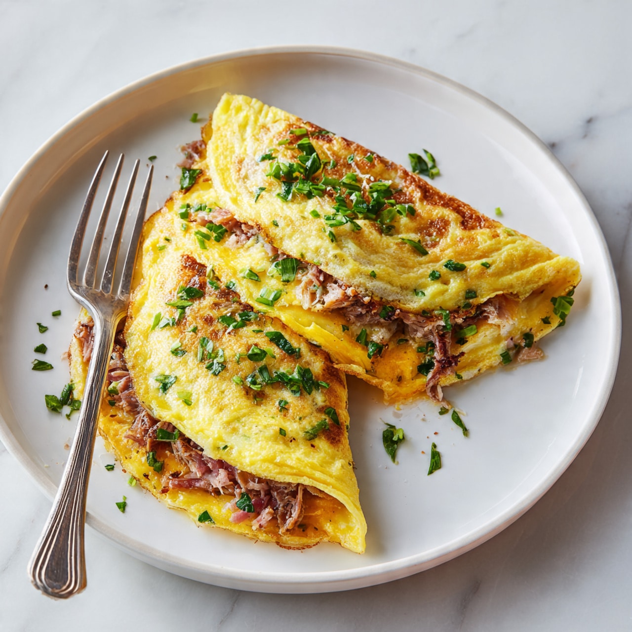 The image shows a folded yellow omelet on a white plate with a fork on the left side. Inside the omelet, there are visible layers of orange cheese, small pieces of meat, and green herbs. The omelet is garnished with chopped green herbs on top. The plate is placed on a white marbled surface with a slight shadow around it. photo taken with an iphone --ar 4:5 --v 7
