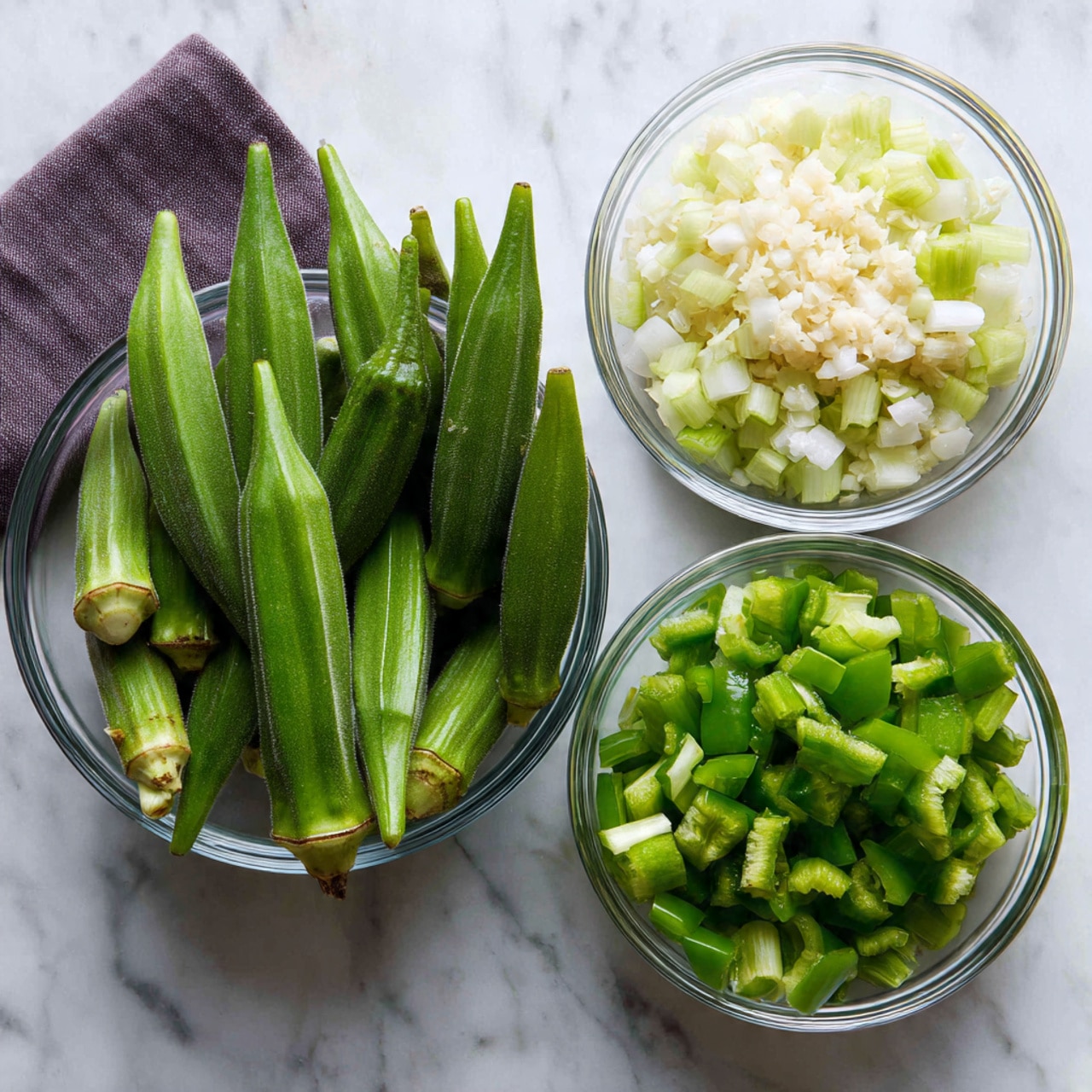 The image shows a clear glass bowl filled with whole fresh green okra pods standing upright in the center on a white marbled surface. Surrounding it are three smaller clear glass bowls arranged in a triangle. The top left bowl contains finely chopped white onions mixed with minced garlic, showing a soft white and light yellow texture. The top right bowl holds chopped celery pieces in light green shades. The bottom bowl is filled with roughly chopped green bell pepper pieces, darker and more vibrant in color. A dark purple cloth is placed in the top left corner of the scene. Photo taken with an iphone --ar 4:5 --v 7